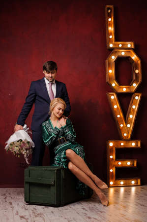 Girl in the dress sitting on the box with her man with bouquet of flowers in his hand on the background light title loveの写真素材
