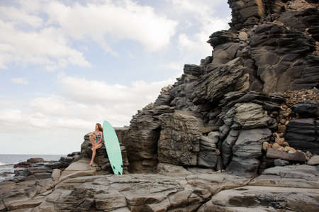 Beautiful sexy girl in the multi colored swimsuit sitting near the surf on the rock on the background of Atlantic ocean and clear skyの写真素材