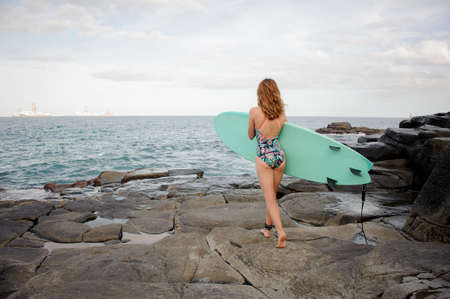 Rear view sporty girl in the multi colored swimsuit walking with the surf on the rocks on the beachの写真素材