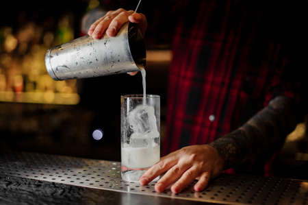 Barman pouring lemonade drink from shaker into a cocktail glass with ice cubes on the barの写真素材