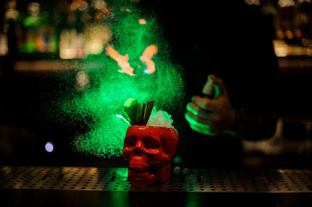 Bartender spraying on the cocktail in the scull cup from the special vaporizer in the green light on...
