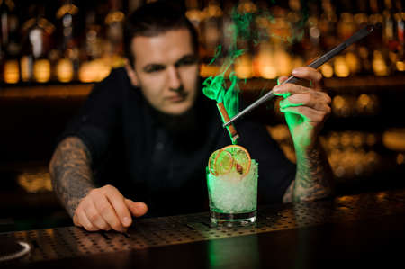 Male bartender adding to the glass with a dried orange and crushed ice aromatic smoked cinnamon with tweezers on the bar counterの写真素材