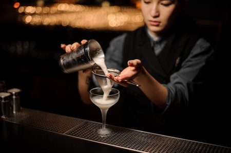 Professional bartender girl pouring a delicious cocktail from the steel shaker through the sieve