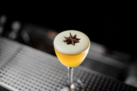 Yellow cocktail with the white foam in the glass decorated with the one anise star standing on the bar counter in the blurred backgroundの写真素材