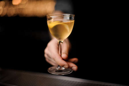 Bartender serving a transparent yellow cocktail in the glass decorated with a lemon zest on the steel bar counter in the blurred dark backgroundの写真素材
