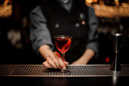 Bartender girl serving a transparent red cocktail in the glass decorated with a lemon zest on the steel bar counter on the steel bar counter in the blurred dark background.の写真素材