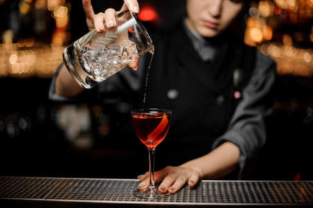 Female bartender pouring an alcohol from the measuring glass cup through the strainer to the cocktail on the bar counter in the dark blurred backgroundの写真素材