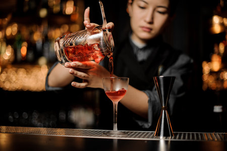 Bartender girl pouring a delicious red cocktail from the measuring glass cup through the strainer on...
