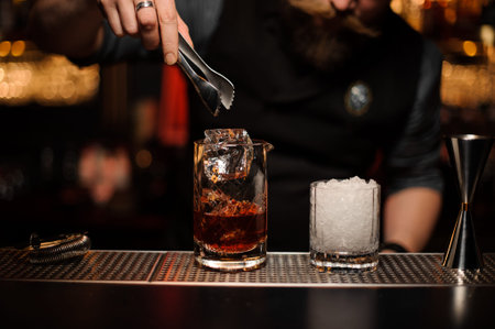 Bartender throwing to a cocktail glass an ice cube with tweezers on the bar counter on the blurred...