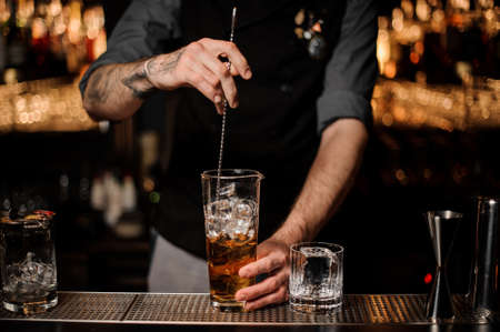 Bartender stirring many ice cubes in the tall glass with a brown cocktail with a spoonの写真素材