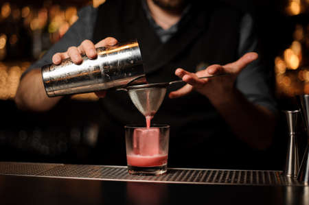Bartender pouring a smooth pink cocktail through the sieve to the glass with one big ice cubeの写真素材