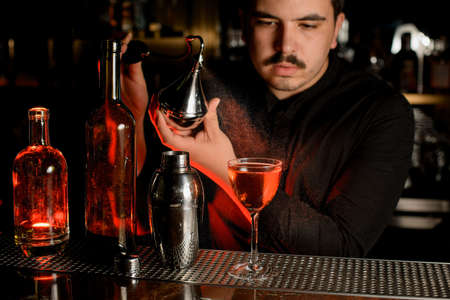 Male bartender with mustache using sprayer to make an alcohol cocktail with bottles and shaker nearbyの写真素材