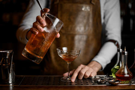 Professional male bartender pouring a delicious brown alcoholic drink from the measuring cup to the cocktail glassの写真素材