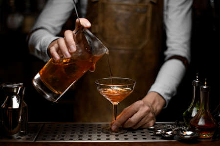 Professional male bartender pouring a brown alcoholic cocktail from the measuring cup to the glassの写真素材