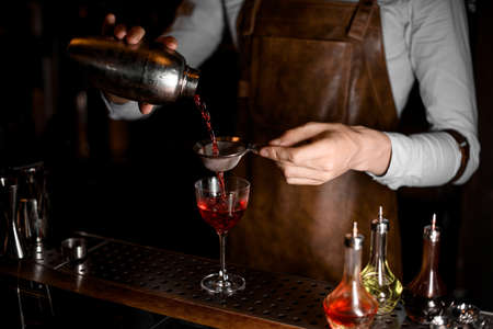Bartender in leather apron pouring a red alcoholic drink from the steel shaker through the sieveの写真素材