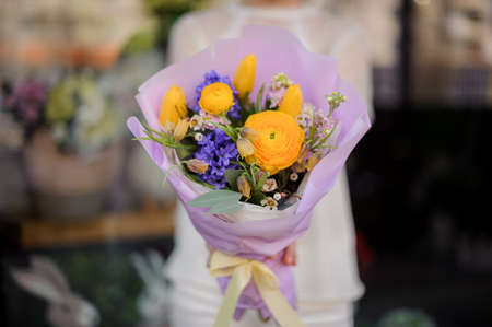 Woman holding a bouquet of yellow peony roses and violet blue hyacinth wrapped in tender color paper decorated with green leaves and branchesの写真素材