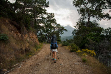 Rear view woman with backpack and hiking sticks walking on the dirt road among the trees and rocksの写真素材