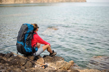Girl sitting on the rocks with hiking backpack and walking sticksの写真素材
