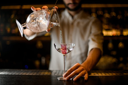 Professional male bartender pouring a cocktail from the measuring cup with a strainer to a glass...