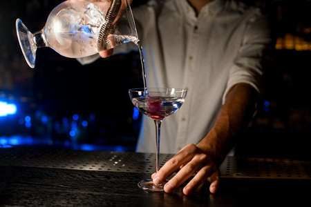 Bartender pouring a cocktail from the measuring cup with a strainer to a glass with pink ice cubeの写真素材