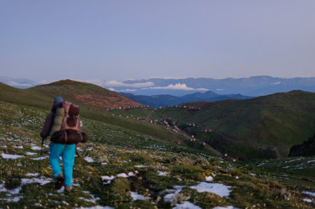 Back view man walking on the field with snow remnants with hiking backpack and sticks