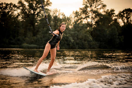 Girl riding on the wakeboard on the river in the sunset holding a rope of the motorboatの写真素材