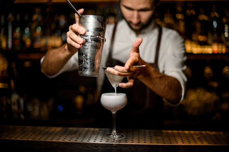Professional male bartender pouring a white cocktail from the steel shaker to the glass through the sieveの写真素材