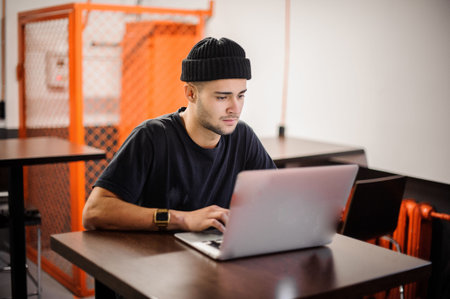 Young hipster man in black hat sitting at the table and using laptop