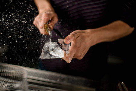 Professional bartender cutting ice with a knife above the empty cocktail glass on the two parts
