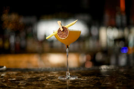Yellow alcoholic cocktail in the glass with a dried lemon and tropical leaf in the clothespin on the bar counterの写真素材