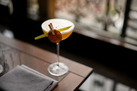 Yellow alcoholic cocktail in the glass with a dried lemon and tropical leaf in the clothespin on the table in the background of windowの写真素材