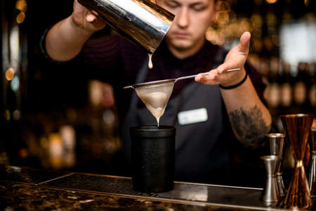 Bartender pouring a alcoholic drink from the steel shaker to the black glass through the sieveの写真素材