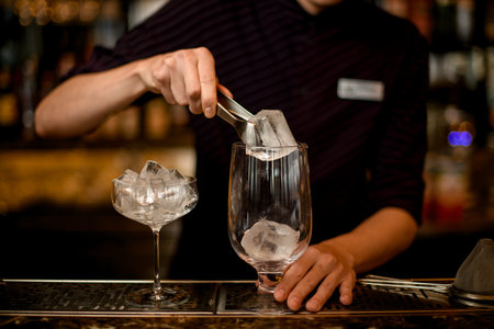 Male bartender putting an ice cube with tweezers to the empty cocktail glassの写真素材