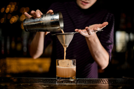Male bartender pouring an alcoholic creamy color drink from the steel shaker to the glass with ice...