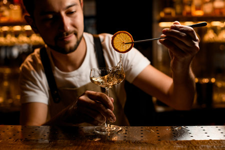 Bartender adding to an alcoholic cocktail a dried lemon slice with tweezers