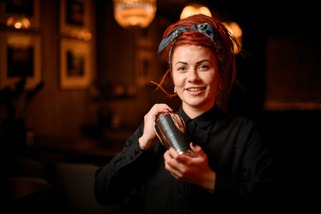 Cute smiling redhead bartender girl with dreadlocks standing in the bar with a steel shakerの写真素材