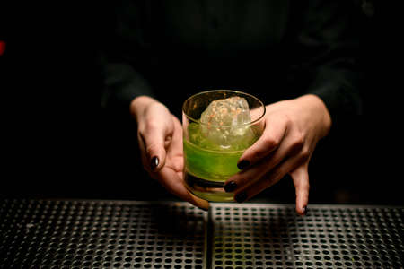 Bartender girl serving green alcoholic cocktail in the glass with a big ice with spices on itの写真素材