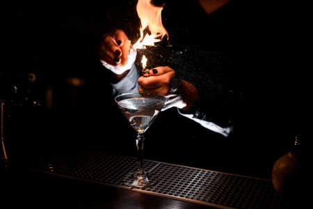 Female bartender sprinkling on a alcoholic drink in the cocktail glass with a orange zest juice and lightening itの写真素材