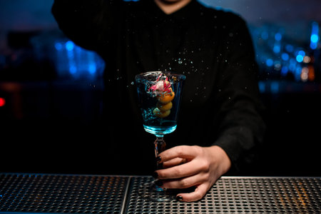 Female bartender serving cocktail in the glass throwing berries and flower on the skewer on it