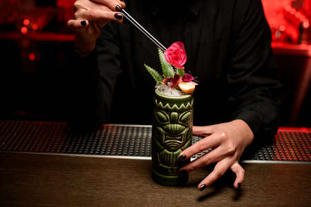 Bartender girl serving alcoholic cocktail in the Tiki mug adding a rose bud with tweezersの写真素材