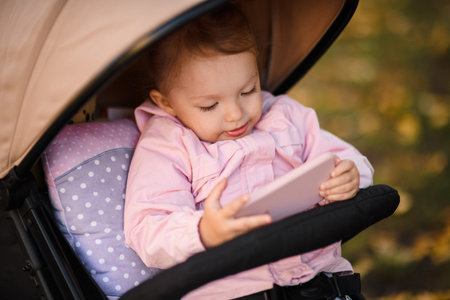 Cute little girl sitting in her baby carriage looking at the phoneの写真素材