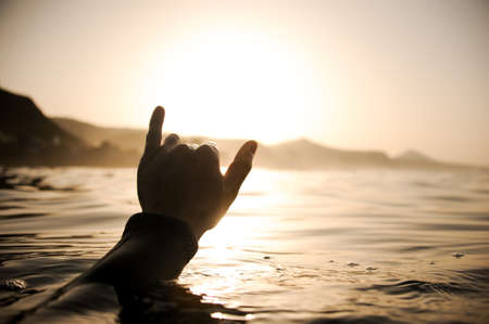 Silhouette man raises his hand out of the water holding up two fingers up in the sunset timeの写真素材
