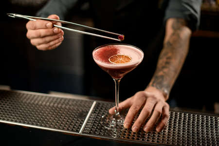 Bartender decorating an alcoholic cocktail in the glass with a dried orange and red spices with tweezers with rose budの写真素材
