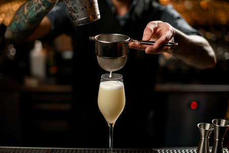 Professional bartender pouring an alcoholic drink from the steel shaker to the glass through the strainer filter on the bar counterの写真素材