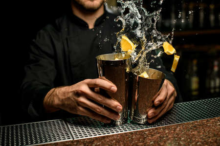 Male bartender holding in hands a steel shakers splashing the cocktail with orange pieces in itの写真素材