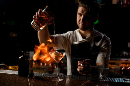 Professional male bartender pouring a brown alcoholic cocktail from the measuring cup to the glass in the fireの写真素材