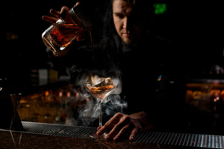 Professional bartender pouring a brown alcoholic drink from the measuring cup to the glass in the...