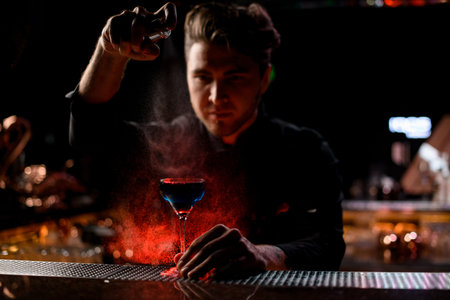 Professional male bartender spraying on the blue alcoholic cocktail with a bitter under red light