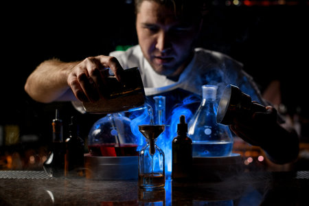 Bartender pouring a transparent alcoholic drink from the steel shaker to the little bottle under...