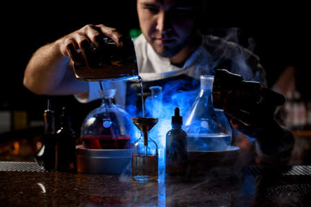 Professional male bartender pouring an alcoholic drink from the steel shaker to the little bottle under blue light on the bar counterの写真素材